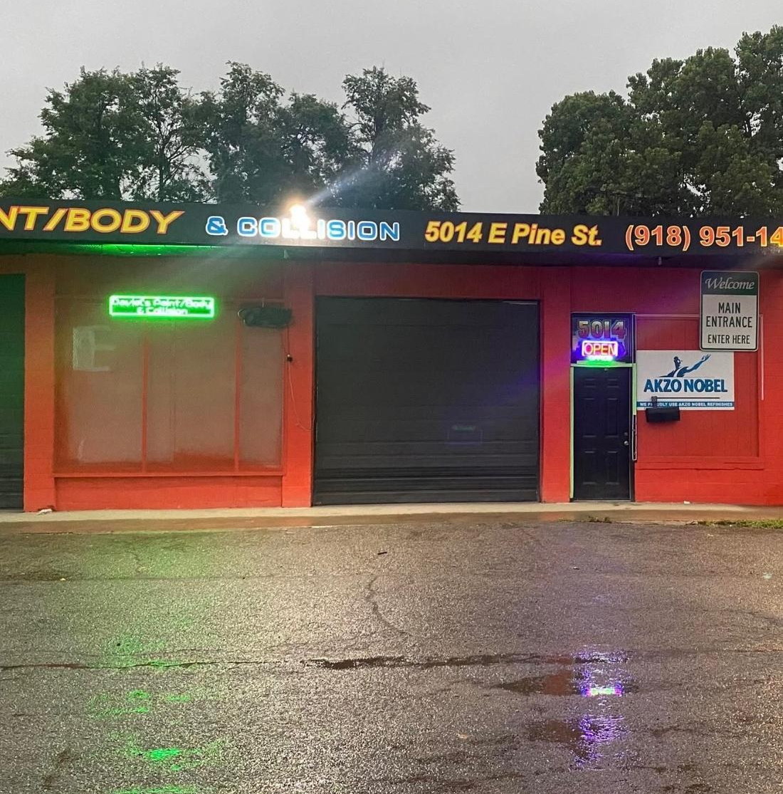 Daviel's Paint, Body & Collision at 5014 E Pine St in Tulsa — neon signs lit at dusk, trees and overcast sky above, rain-slick parking lot in front